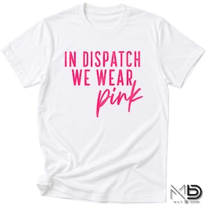 May include: A white short-sleeved t-shirt featuring the text IN DISPATCH WE WEAR pink printed in a bright pink colour on the front. The shirt has a crew neckline and a standard fit, with the text displayed in a mix of block and script fonts.
