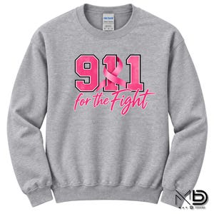 Op de afbeelding: Een grijs gemêleerd sweatshirt met ronde hals en de tekst 911 in grote roze blokletters met een zwarte omlijning. Een roze bewustzijnslint is verwerkt in het cijfer 1. Daaronder staat de tekst for the Fight in een roze cursief lettertype.