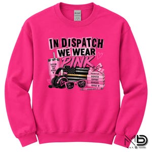May include: A bright pink long-sleeve sweatshirt featuring a graphic print with the text In Dispatch We Wear Pink. The design includes a pink tumbler, black headphones, a flag, pink sunglasses, a stack of books, and a pen with various text labels.