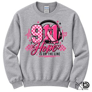 Könnte beinhalten: Ein graumeliertes Sweatshirt mit Rundhalsausschnitt und einer Grafik, die die Zahl 911 in einem rosa Karomuster, eine rosa Schleife und einen schwarzen Kopfhörer zeigt. Der Text lautet 911 Hope is on the line, verziert mit rosa Herzen und Sternen.