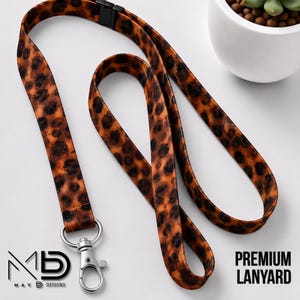 May include: A fabric lanyard featuring a brown and black leopard print pattern. The lanyard includes a black plastic breakaway clasp at the top and a silver-toned metal swivel lobster claw clasp at the bottom for attaching items securely.
