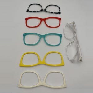 Op de afbeelding: Een assortiment brillen en monturen in verschillende kleuren. De monturen omvatten zwart-wit gespikkeld, rood, teal, geel en wit. Er is ook een bril van helder plastic aanwezig, die een verscheidenheid aan brilopties biedt.