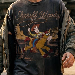 K&ouml;nnte beinhalten: Dunkelgraues Langarm-T-Shirt mit Sheriff Woody und Pferde-Grafik. Die Grafik zeigt Woody, der mit einem Lasso auf einem Pferd reitet. Das Shirt hat einen Vintage-Look.