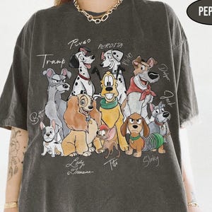 以下が含まれることがあります： ポング、ペルディタ、レディなど、さまざまな犬のキャラクターのカラフルな漫画グラフィックが特徴の濃い灰色のTシャツ。「Pepper」の文字が右上隅に印刷されています。