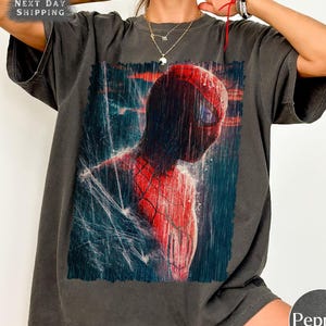 Pu&ograve; includere: T-shirt grigio scuro con una vivace immagine di Spider-Man. Il supereroe &egrave; raffigurato nella sua iconica tuta rossa e nera, sullo sfondo di pioggia e dettagli di ragnatela. La maglietta ha anche il testo "NEXT DAY SHIPPING".