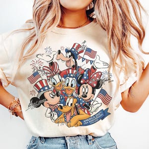 May include: Cream-coloured t-shirt featuring a graphic of Mickey Mouse, Minnie Mouse, Donald Duck, Daisy Duck, Goofy, and Pluto, all in patriotic attire with American flags and fireworks. The shirt commemorates the 250th USA Anniversary.