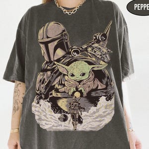May include: Dark gray t-shirt featuring a graphic of The Mandalorian, Grogu, and a spaceship. The design includes the text "PEPPER" in a black oval. The shirt has short sleeves and a relaxed fit.