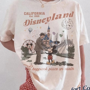 May include: Ivory-colored t-shirt with a Disneyland graphic. The graphic features Mickey Mouse, a man in a suit, and iconic park imagery. Text includes "California Disneyland Est. 1955" and "The happiest place on earth."