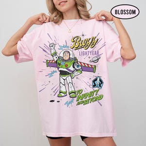 May include: A light pink t-shirt featuring a graphic of Buzz Lightyear from Toy Story. The design includes the text "Buzz Lightyear" and "To Infinity and Beyond" with a space-themed background. The shirt is oversized.