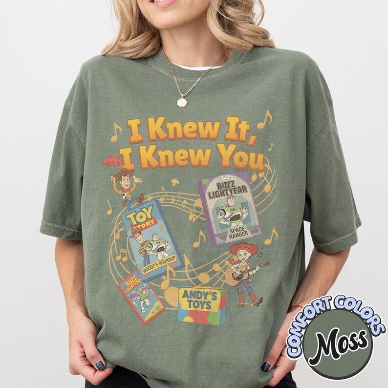 K&ouml;nnte beinhalten: Ein olivgr&uuml;nes Kurzarm-T-Shirt mit einem Grafikdruck und dem Text I Knew It, I Knew You. Das Design zeigt Illustrationen von Toy Story-Figuren, VHS-H&uuml;llen, Musiknoten und eine Schachtel mit der Aufschrift Andy's Toys.