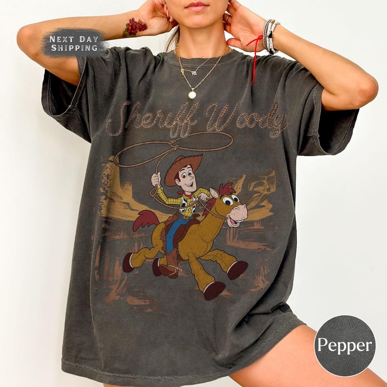 K&ouml;nnte beinhalten: Dunkelgraues T-Shirt mit einem Used-Look-Grafik von Sheriff Woody, der auf einem Pferd mit einem Lasso reitet. Der Text "Sheriff Woody" ist oben aufgestickt. Das Shirt hat einen Vintage-Look.