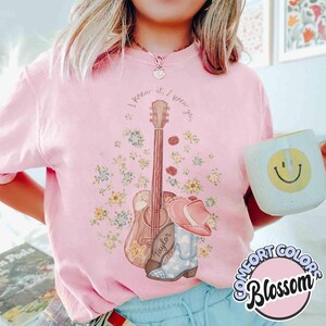 May include: A light pink t-shirt featuring a graphic print of an acoustic guitar, a cowboy boot, and colorful flowers. The text "I knew you" is arched above the guitar. The shirt is paired with a light blue mug with a yellow smiley face.