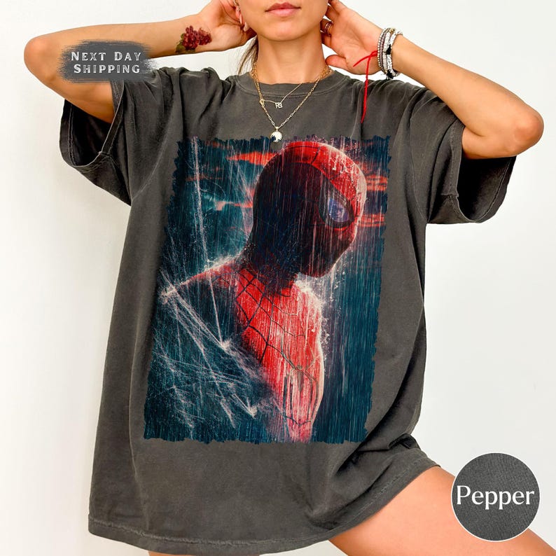 Pu&ograve; includere: T-shirt grigio scuro con una vivace immagine di Spider-Man. Il supereroe &egrave; raffigurato nella sua iconica tuta rossa e nera, sullo sfondo di pioggia e dettagli di ragnatela. La maglietta ha anche il testo "NEXT DAY SHIPPING".