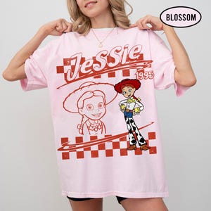 May include: A light pink t-shirt featuring a graphic of the character Jessie from the movie Toy Story. The design includes the name "Jessie" in a stylized font, the year "1995", and a red checkered pattern.