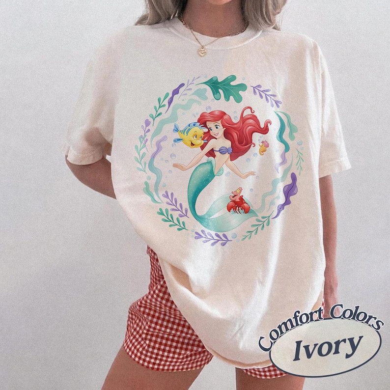 May include: Ivory-colored t-shirt featuring a circular graphic of a mermaid with red hair, a teal tail, and a yellow fish. The design is surrounded by aquatic plants and other sea creatures. The shirt is labeled "Comfort Colors Ivory".