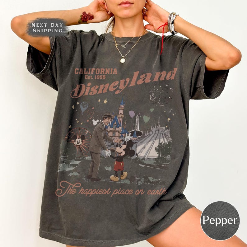 Puede incluir: Camiseta gris oscuro con un gr&aacute;fico de Disneyland. El dise&ntilde;o presenta una ilustraci&oacute;n vintage de Mickey Mouse, un hombre y el castillo de Disneyland. El texto incluye "CALIFORNIA Disneyland Est. 1955" y "The happiest place on earth."