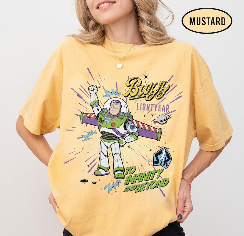 May include: Mustard-colored t-shirt featuring a graphic of Buzz Lightyear from Toy Story. The design includes the words "Buzz Lightyear" and "To Infinity and Beyond." The shirt has short sleeves and a crew neck.
