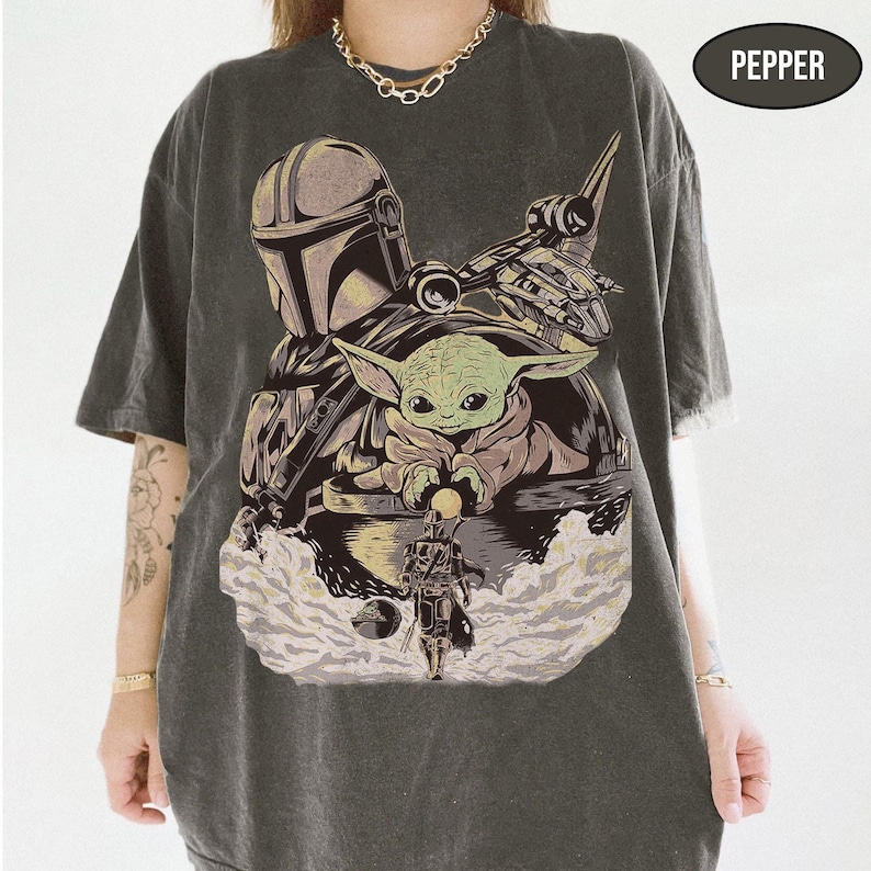 May include: Dark gray t-shirt featuring a graphic of The Mandalorian, Grogu, and a spaceship. The design includes the text "PEPPER" in a black oval. The shirt has short sleeves and a relaxed fit.