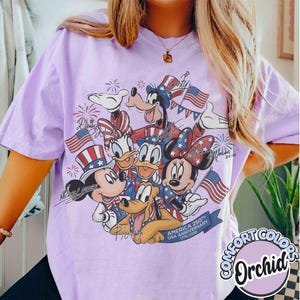 May include: A lilac Comfort Colors t-shirt with a graphic of Disney characters Mickey Mouse, Minnie Mouse, Donald Duck, Daisy Duck, Goofy, and Pluto in patriotic outfits. The design includes American flags and fireworks, celebrating the USA's 250th anniversary.