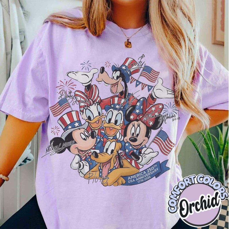 May include: A lilac Comfort Colors t-shirt with a graphic of Disney characters Mickey Mouse, Minnie Mouse, Donald Duck, Daisy Duck, Goofy, and Pluto in patriotic outfits. The design includes American flags and fireworks, celebrating the USA's 250th anniversary.