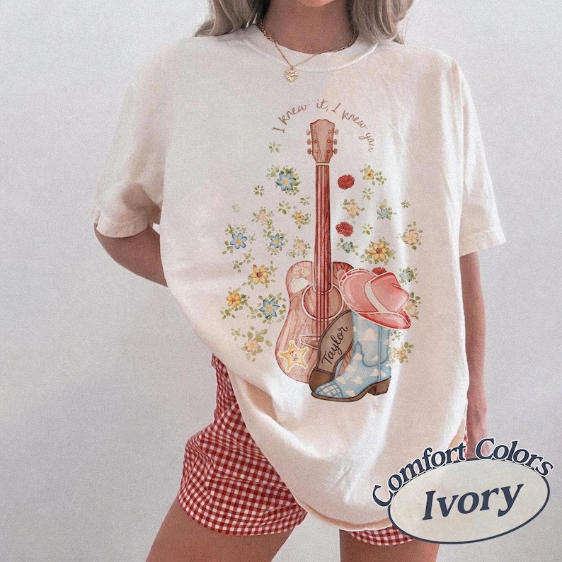May include: An ivory t-shirt featuring a graphic print of an acoustic guitar, a pink cowboy hat, and a light blue cowboy boot with the name Taylor written on it. The design is surrounded by small colorful flowers and the text I knew it, I knew you.