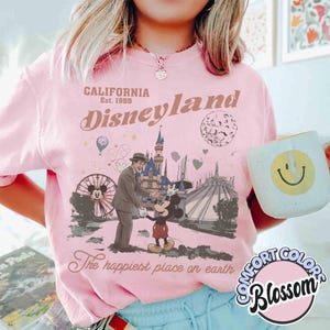 Puede incluir: Camiseta rosa p&aacute;lido de Disneyland con un dise&ntilde;o vintage que muestra a Mickey Mouse, un hombre y atracciones del parque. El texto dice "CALIFORNIA Disneyland Est. 1955" y "The happiest place on earth". Se sostiene una taza blanca con una carita sonriente amarilla.