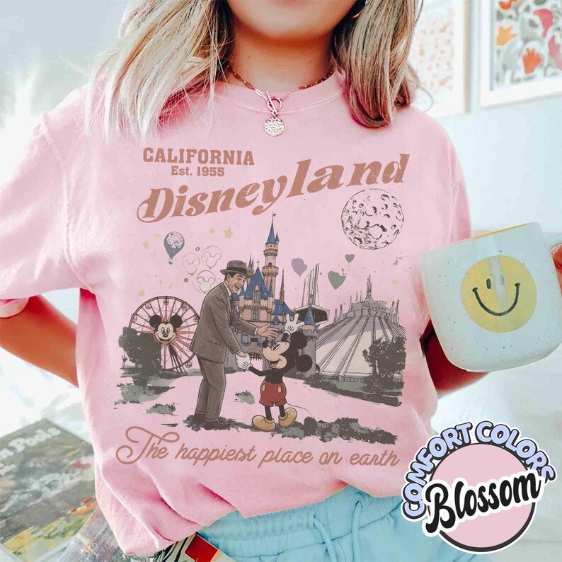 Puede incluir: Camiseta rosa p&aacute;lido de Disneyland con un dise&ntilde;o vintage que muestra a Mickey Mouse, un hombre y atracciones del parque. El texto dice "CALIFORNIA Disneyland Est. 1955" y "The happiest place on earth". Se sostiene una taza blanca con una carita sonriente amarilla.