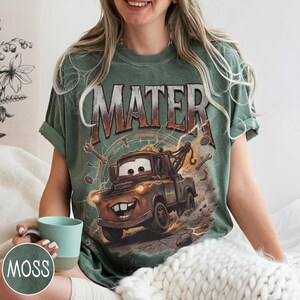 Puede incluir: Camiseta verde con una imagen de dibujos animados de Mate de la pel&iacute;cula Cars. El gr&aacute;fico incluye el nombre "MATER" encima del personaje. La camiseta tiene un aspecto vintage y desgastado.