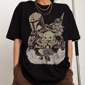 May include: Black t-shirt featuring a graphic of The Mandalorian, Grogu, and a spaceship. The design includes a helmeted figure, a green-skinned baby, and a detailed spacecraft, all set against a cloudy background.