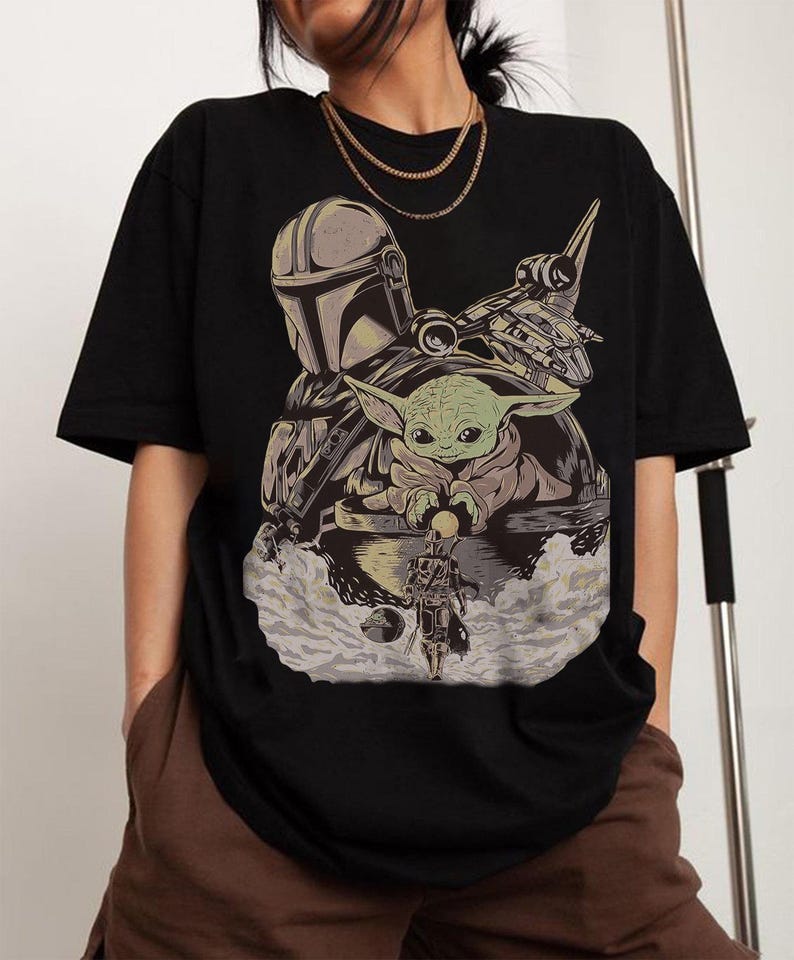 May include: Black t-shirt featuring a graphic of The Mandalorian, Grogu, and a spaceship. The design includes a helmeted figure, a green-skinned baby, and a detailed spacecraft, all set against a cloudy background.