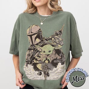 May include: Olive green t-shirt featuring a graphic of The Mandalorian, Grogu, and a spaceship. The design is in shades of gray, tan, and green. The shirt has a crew neck and short sleeves. The words "COMFORT COLORS" and "Moss" are printed on the lower right.