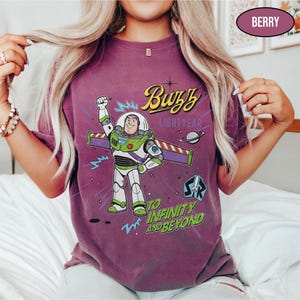 May include: Berry-colored t-shirt featuring a graphic of Buzz Lightyear with the text "Buzz Lightyear" and "To Infinity and Beyond." The design includes a cartoon image of Buzz Lightyear in his space suit. The shirt has short sleeves.