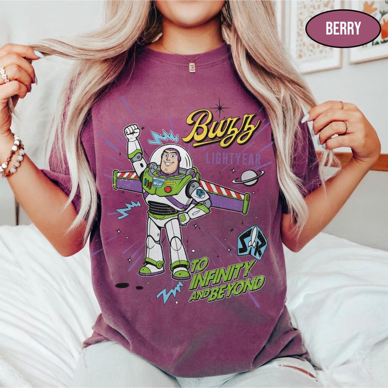 May include: Berry-colored t-shirt featuring a graphic of Buzz Lightyear with the text "Buzz Lightyear" and "To Infinity and Beyond." The design includes a cartoon image of Buzz Lightyear in his space suit. The shirt has short sleeves.