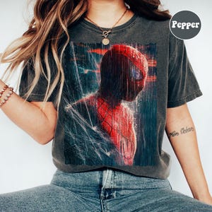 Pu&ograve; includere: T-shirt grigio scuro con una vivace illustrazione di Spider-Man in una scena piovosa. Il supereroe &egrave; raffigurato nel suo iconico costume rosso e blu, con gocce d'acqua e dettagli di ragnatela. La maglietta ha un aspetto vintage e usurato.
