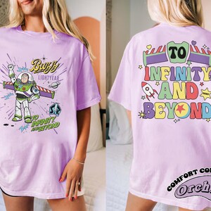 Op de afbeelding: Lila T-shirt met een Buzz Lightyear-afbeelding en de tekst "Buzz Lightyear" en "To Infinity and Beyond". De achterkant van het shirt toont "To Infinity and Beyond" in kleurrijke letters, een raket en sterren. Het shirt is een Comfort Colors Orchid.