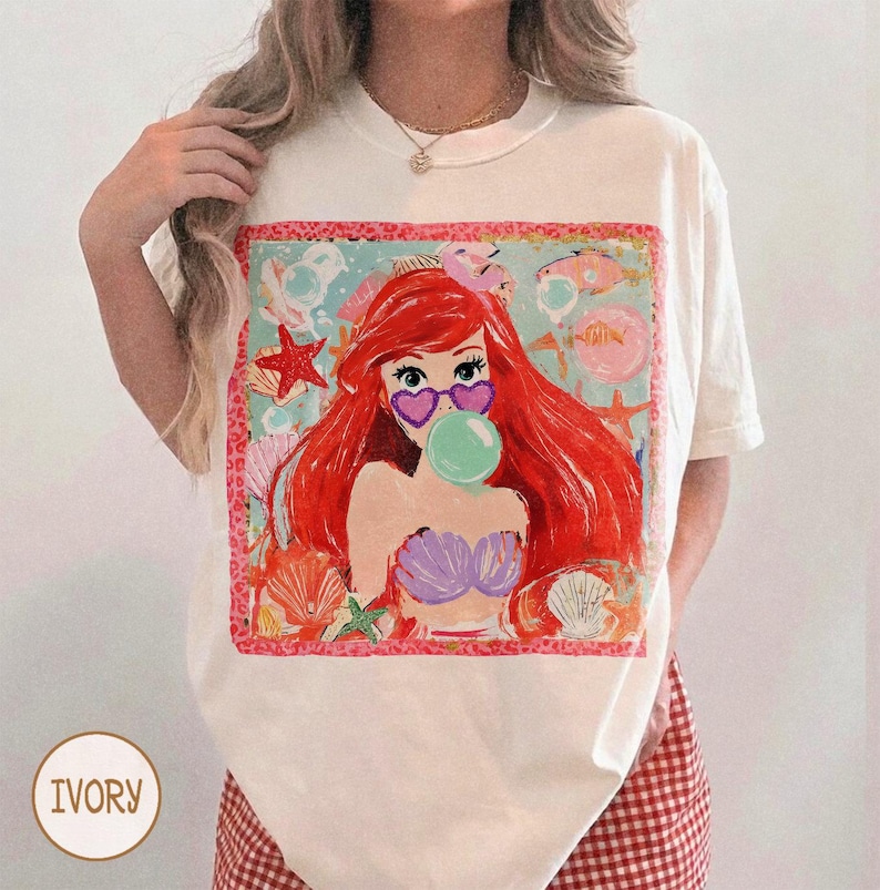 May include: Ivory-colored t-shirt featuring a colorful graphic of a cartoon mermaid with red hair, heart-shaped sunglasses, and a bubble. The graphic includes seashells, starfish, and fish. The shirt has a round neckline and short sleeves.