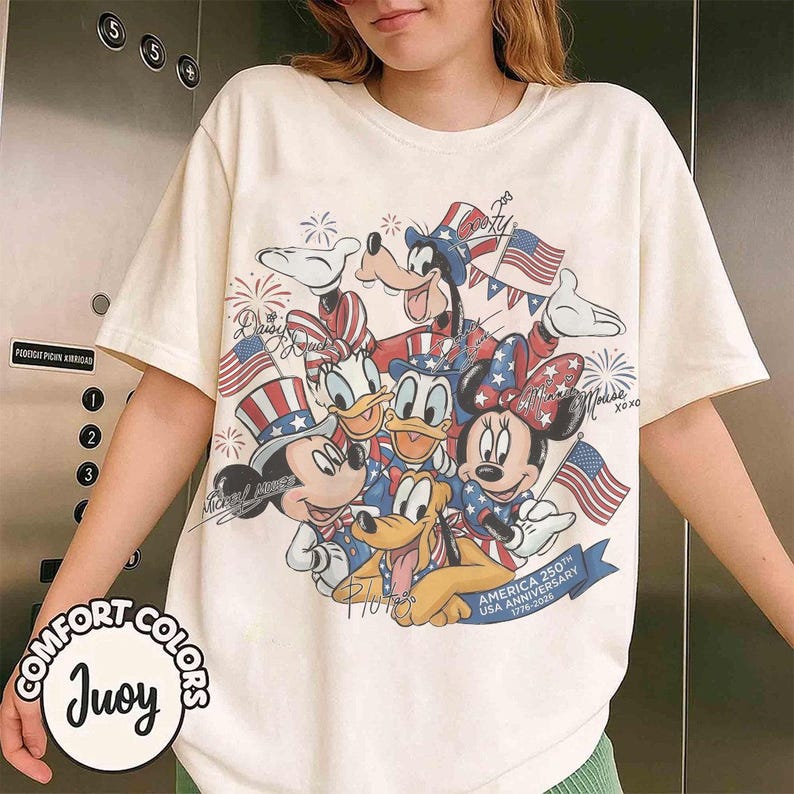 May include: Cream-coloured t-shirt with a graphic of Disney characters, including Mickey and Minnie Mouse, Donald Duck, and Goofy, celebrating the USA's 250th anniversary. The design incorporates American flags and patriotic elements.