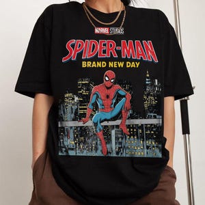 May include: Black t-shirt with the Marvel Studios Spider-Man logo and the text "BRAND NEW DAY". The design depicts Spider-Man seated on a rooftop, with a city skyline in the background.