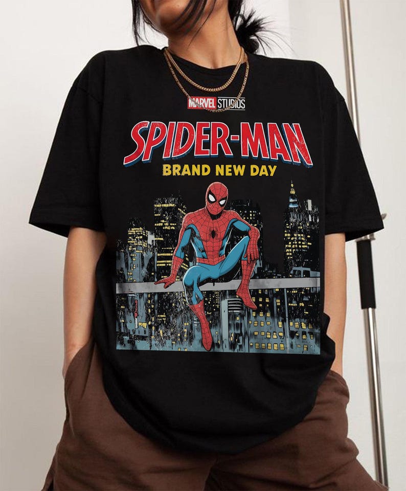 May include: Black t-shirt with the Marvel Studios Spider-Man logo and the text "BRAND NEW DAY". The design depicts Spider-Man seated on a rooftop, with a city skyline in the background.