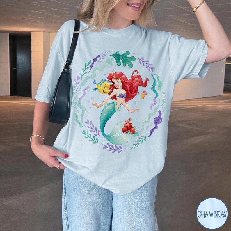 May include: Pale blue t-shirt with a colourful illustration of Ariel from The Little Mermaid. The design features Ariel with red hair, a turquoise tail, and her friends Flounder and Sebastian, surrounded by a floral wreath. The shirt has a relaxed fit.