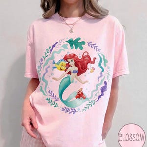 May include: A pale pink t-shirt with a cartoon illustration of a mermaid with red hair and a teal tail. The design includes a yellow fish, a red crab, and aquatic plants. The shirt has short sleeves and a round neckline. The word "Blossom" is in a circle.