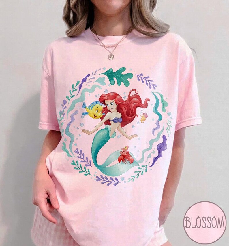 May include: A pale pink t-shirt with a cartoon illustration of a mermaid with red hair and a teal tail. The design includes a yellow fish, a red crab, and aquatic plants. The shirt has short sleeves and a round neckline. The word "Blossom" is in a circle.