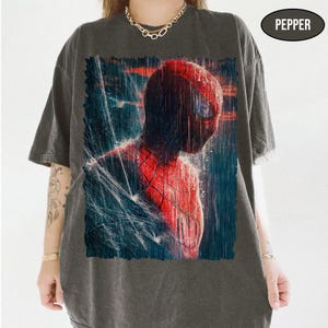 Pu&ograve; includere: T-shirt grigio scuro con una grafica di Spider-Man in una scena piovosa. Il costume rosso e nero del supereroe &egrave; dettagliato, con un focus sulla sua maschera. La parola "PEPPER" &egrave; stampata in alto a destra sulla maglietta.