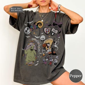 May include: Dark gray t-shirt featuring a collage of characters and imagery from "The Nightmare Before Christmas." The design includes Jack Skellington, Sally, Oogie Boogie, and other elements. The shirt has the text "NIGHTMARE" and the color "Pepper".