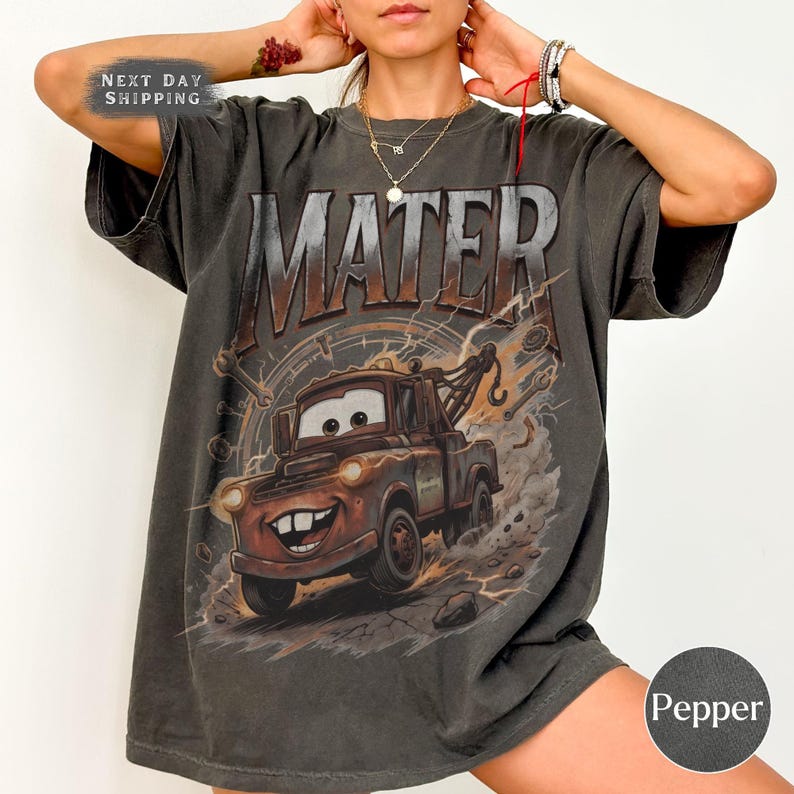 Puede incluir: Camiseta gris oscuro con una imagen de dibujos animados de Mate de la pel&iacute;cula Cars. La palabra "MATER" est&aacute; impresa en la parte superior con una fuente desgastada. La imagen muestra a Mate con una cara sonriente y caracter&iacute;sticas de gr&uacute;a. La camiseta tiene un aspecto vintage.