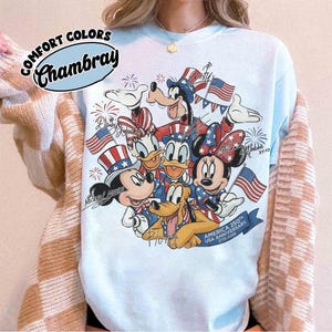 May include: Pale blue Comfort Colors Chambray sweatshirt with a patriotic design featuring Mickey Mouse, Minnie Mouse, Donald Duck, Daisy Duck, Goofy, and Pluto. The graphic includes American flags and fireworks, celebrating the USA's 250th Anniversary.