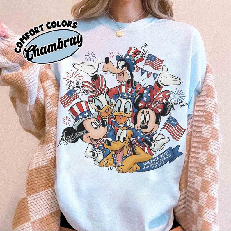 May include: Pale blue Comfort Colors Chambray sweatshirt with a patriotic design featuring Mickey Mouse, Minnie Mouse, Donald Duck, Daisy Duck, Goofy, and Pluto. The graphic includes American flags and fireworks, celebrating the USA's 250th Anniversary.