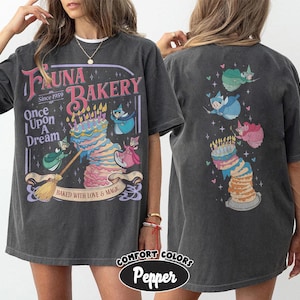 May include: A charcoal gray t-shirt featuring a graphic print of a multi-layered cake with candles, three small fairy figures, and text that reads Fauna Bakery Since 1959 Once Upon A Dream Baked With Love & Magic on the front and back.