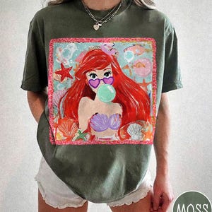 May include: Moss green t-shirt featuring a colorful graphic of a mermaid with red hair, heart-shaped sunglasses, and a bubble. The graphic includes seashells, starfish, and fish. The shirt is a relaxed fit.