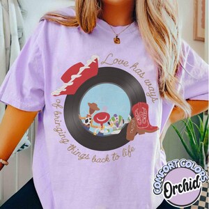 May include: A light purple t-shirt featuring a graphic of a black vinyl record with a central illustration of three cartoon characters, a red cowboy hat, and a red cowboy boot. The text reads, Love has ways of bringing things back to life.